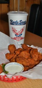 A basket of fried chicken wings with a side of ranch dip, vegetables, and an Uncle Sam’s branded drink cup on a table.