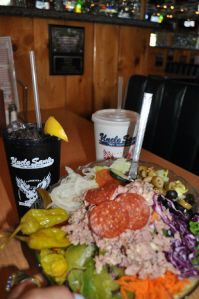 A salad topped with pepperoni, tuna, and peppers, alongside two drinks at a table inside a restaurant.