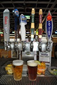 Six beer tap handles line a bar counter above two glasses of beer, one light and one dark, with citrus wedges.