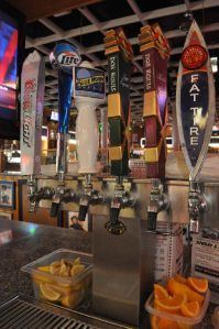 Six beer taps lined up on a bar with bowls of sliced lemons and oranges sitting on the counter underneath them.