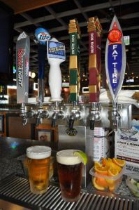 Six beer tap handles line a bar counter, with two filled glasses of beer and a bowl of orange slices in the foreground.