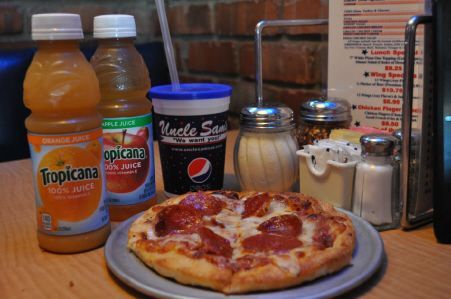 A small pepperoni pizza on a plate next to two bottles of Tropicana juice, a soda cup, and condiments on a restaurant table.