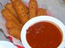 A basket containing several golden-brown fried mozzarella sticks next to a white bowl of red marinara dipping sauce.