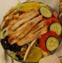 A chicken salad with grilled slices, tomato, cucumber, black beans, and pickles in a glass bowl.