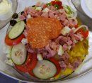 A plated meal featuring mashed plantains topped with diced ham, fresh cucumber and tomato slices, and olives.