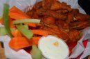 A basket of fried chicken wings served with carrot sticks, celery, and a side of white dipping sauce on parchment paper.