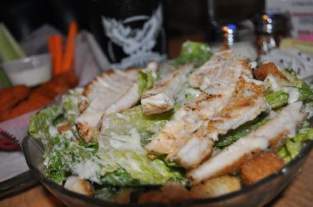 A chicken Caesar salad in a glass bowl, with side dishes and a glass in the blurred background.
