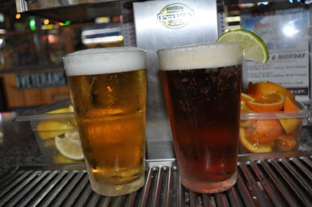 Two glasses of beer, one light and one dark, sit on a bar drip tray with lemon and orange wedges nearby.