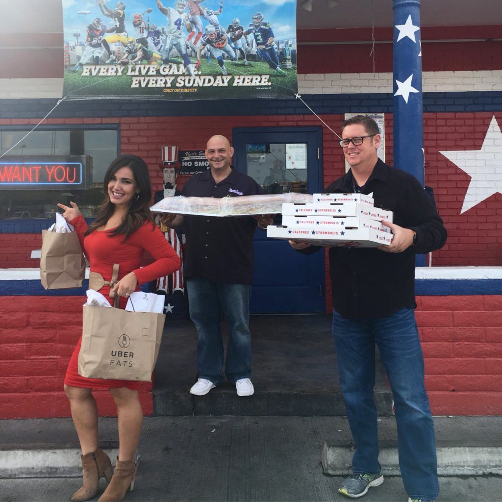 Three people stand outside a restaurant with red and blue brick walls, holding takeout bags and stacks of pizza boxes.