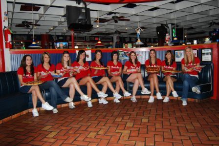 Ten waitresses in red shirts and jeans sit in a row on blue booth seating, holding pizzas inside a restaurant.