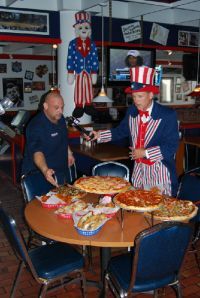 A person in an Uncle Sam costume stands with a man at a table laden with several pizzas in a restaurant setting.