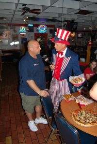 A person in an Uncle Sam costume holds a microphone and a food basket while talking to a man in a restaurant.