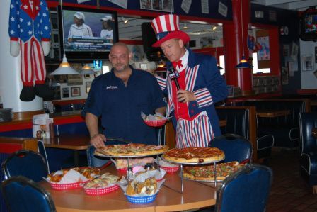 A person in an Uncle Sam costume interviews a man standing by a table filled with various pizzas in a themed restaurant.
