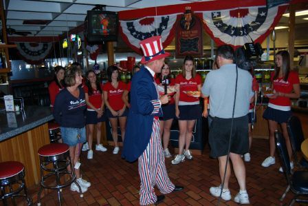A person dressed as Uncle Sam stands in a restaurant with staff wearing matching red shirts, speaking into a microphone.