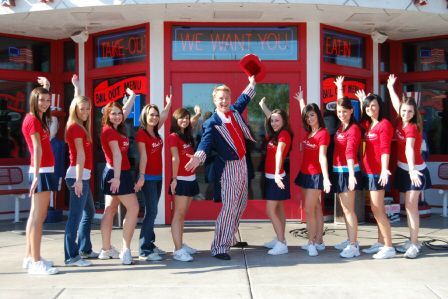 A man in a patriotic costume stands with ten people in red shirts and navy skirts in front of a building saying WE WANT YOU.