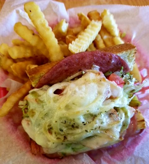 A toasted sandwich with melted cheese, cured meat, and vegetables served with a side of crinkle-cut fries in a paper liner.