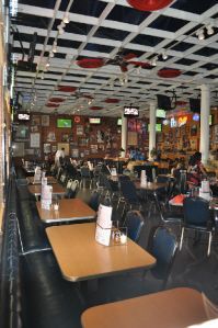 Empty dining tables with chairs arranged in a casual, sports-themed restaurant with a grid ceiling and wall TVs.