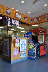 A shop counter with posters, an American flag, and an arcade cabinet in a room with wood-paneled walls and ceiling lights.