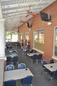 Outdoor dining patio with brick walls, blue chairs, tables, ceiling fans, and a mounted television.