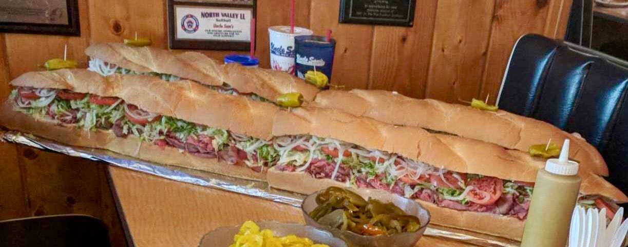 A massive sub sandwich sits on a table in a restaurant with wood-paneled walls, beside condiments and drink cups.