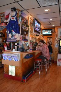 A person sits at a bar counter inside a dimly lit establishment decorated with sports banners, a plush toy, and televisions.