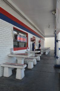 Outdoor seating area with white stone tables and benches against a white brick wall featuring red and blue stripes.