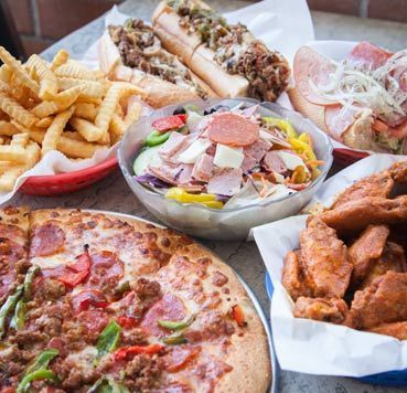 A spread of fast food including pizza, chicken wings, french fries, two sub sandwiches, and an antipasto salad.