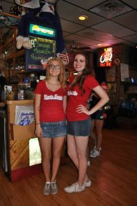 Two people in red shirts and denim skirts stand smiling in a bar with wooden walls and Pepsi signage.