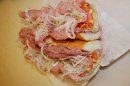 A top-down view of a sandwich on a white wrapper, featuring sliced deli meat, shredded onions, and a red spread.