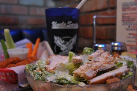 A salad with sliced grilled chicken in a glass bowl, next to a basket of celery, carrots, and a dark blue drink cup.