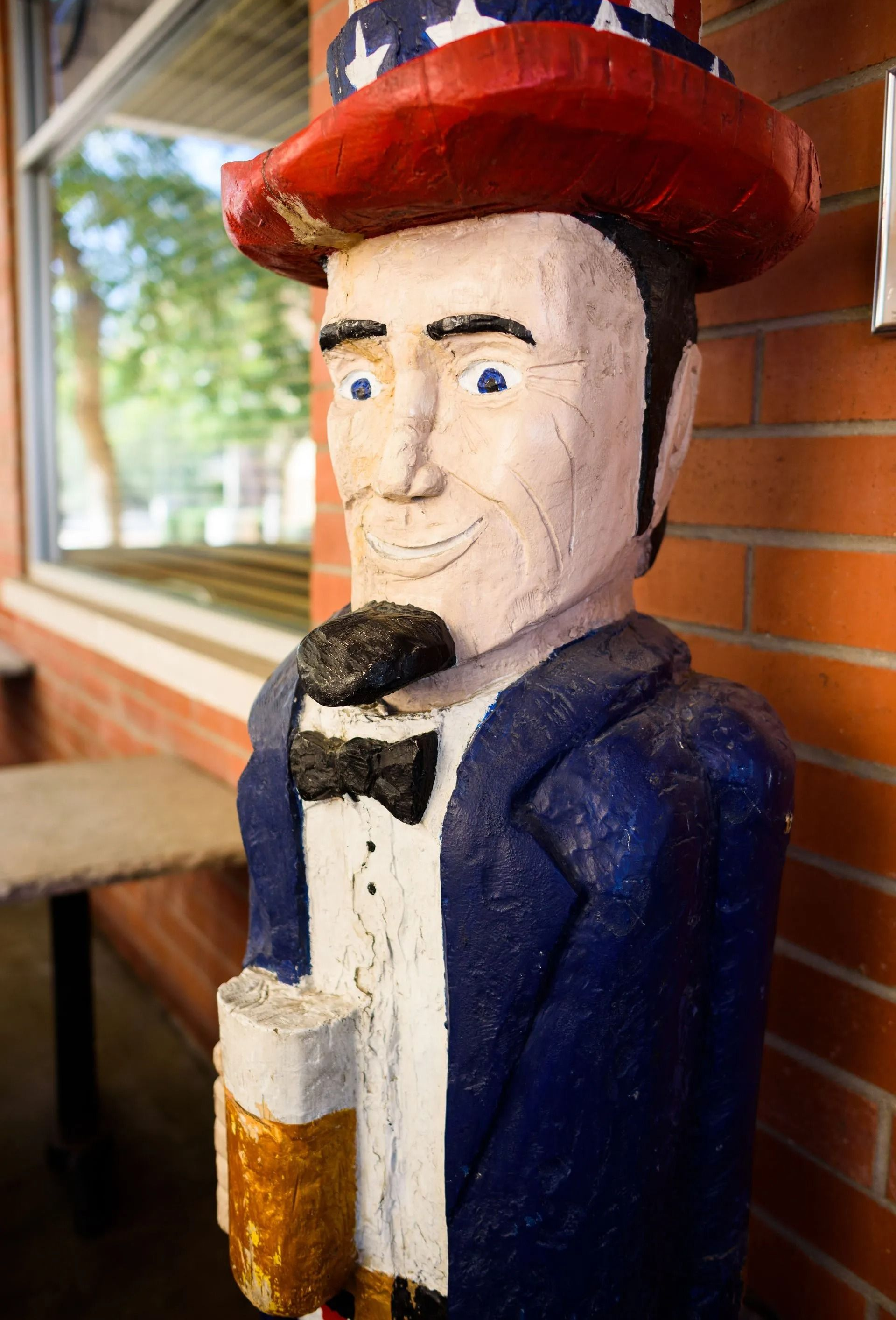 A carved wooden statue of a figure wearing a tall, star-spangled top hat, blue blazer, and bow tie, standing by a wall.