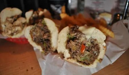 A close-up view of several cheesesteak sandwiches in rolls, served on white parchment paper with a side of fries.