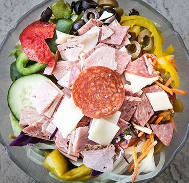 A chopped salad in a glass bowl, featuring sliced deli meats, pepperoni, cheese, cucumbers, peppers, and olives.