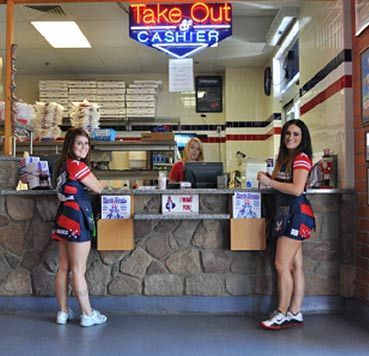 Two employees in matching red and blue uniforms stand at a pizza shop counter, while a third works in the background.