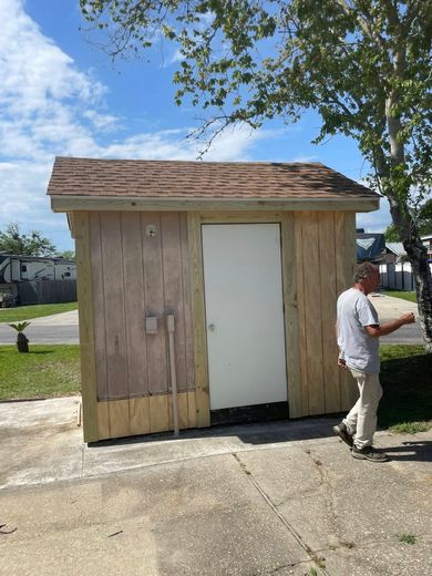 A person stands beside a small, wooden storage shed with a white door and shingled roof, located on a paved lot.