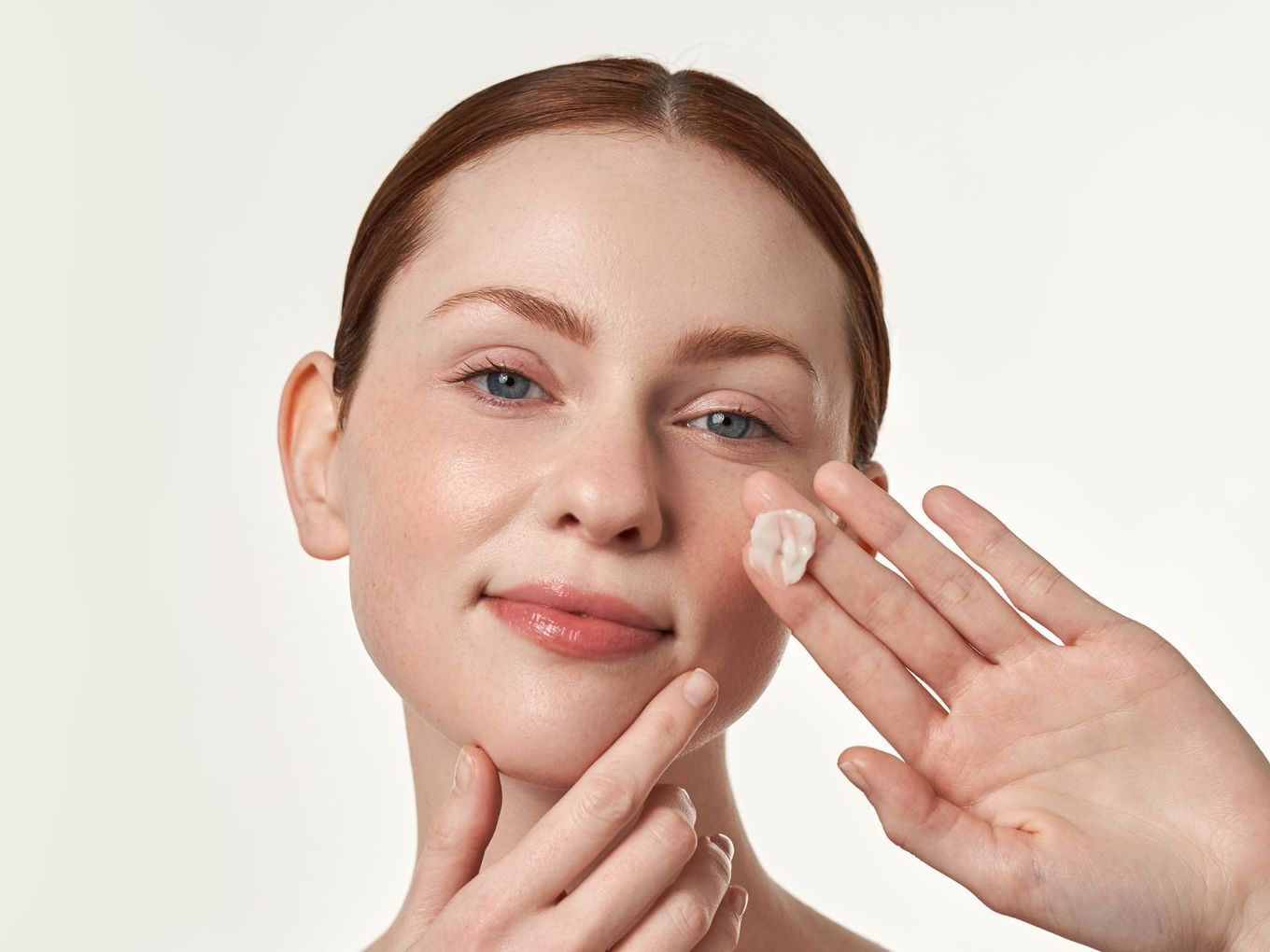 Woman with fair skin applying cream to her face, smiling with red hair, and light background.