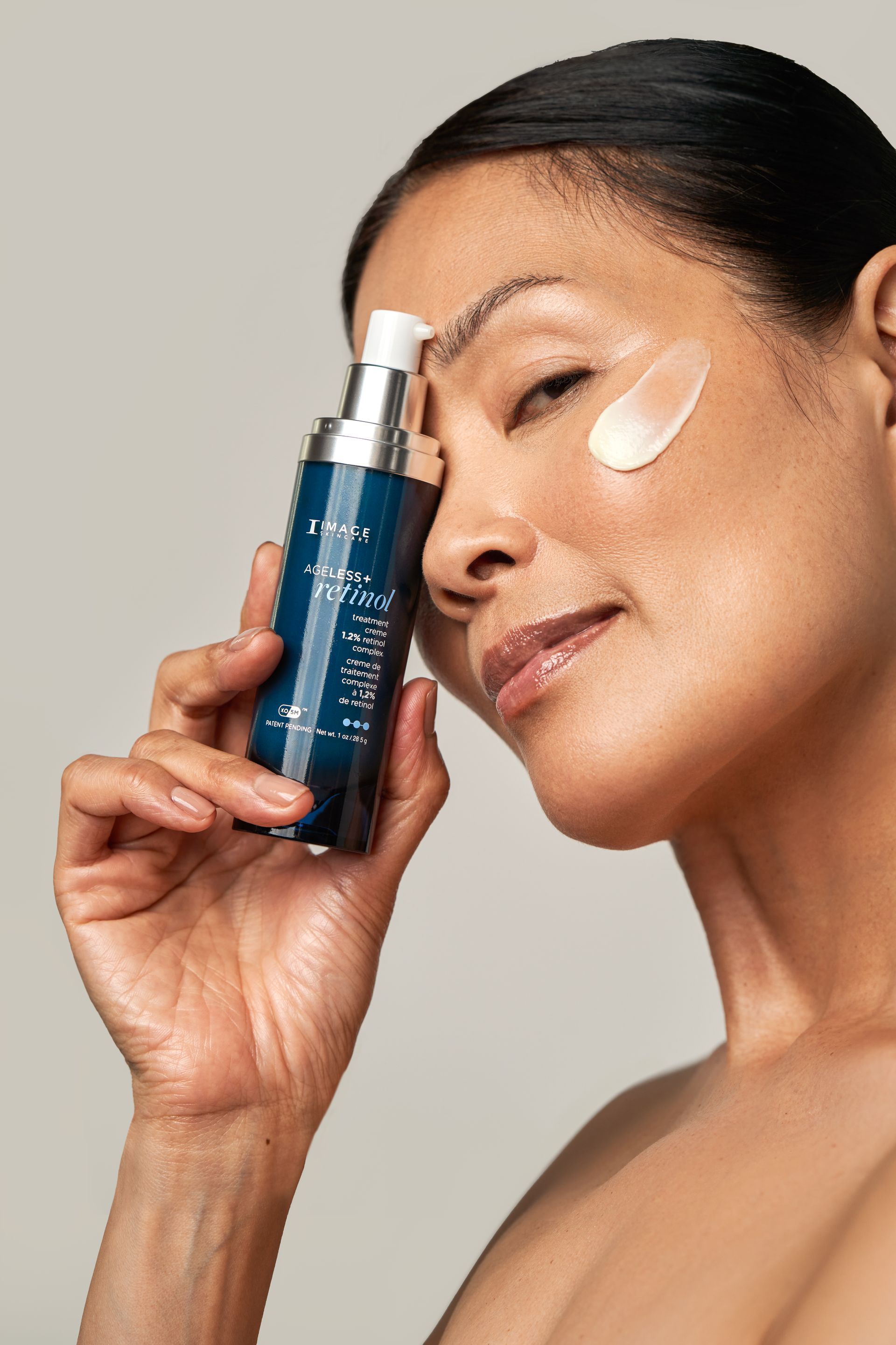 Woman holding skincare bottle, cream on face, soft lighting.