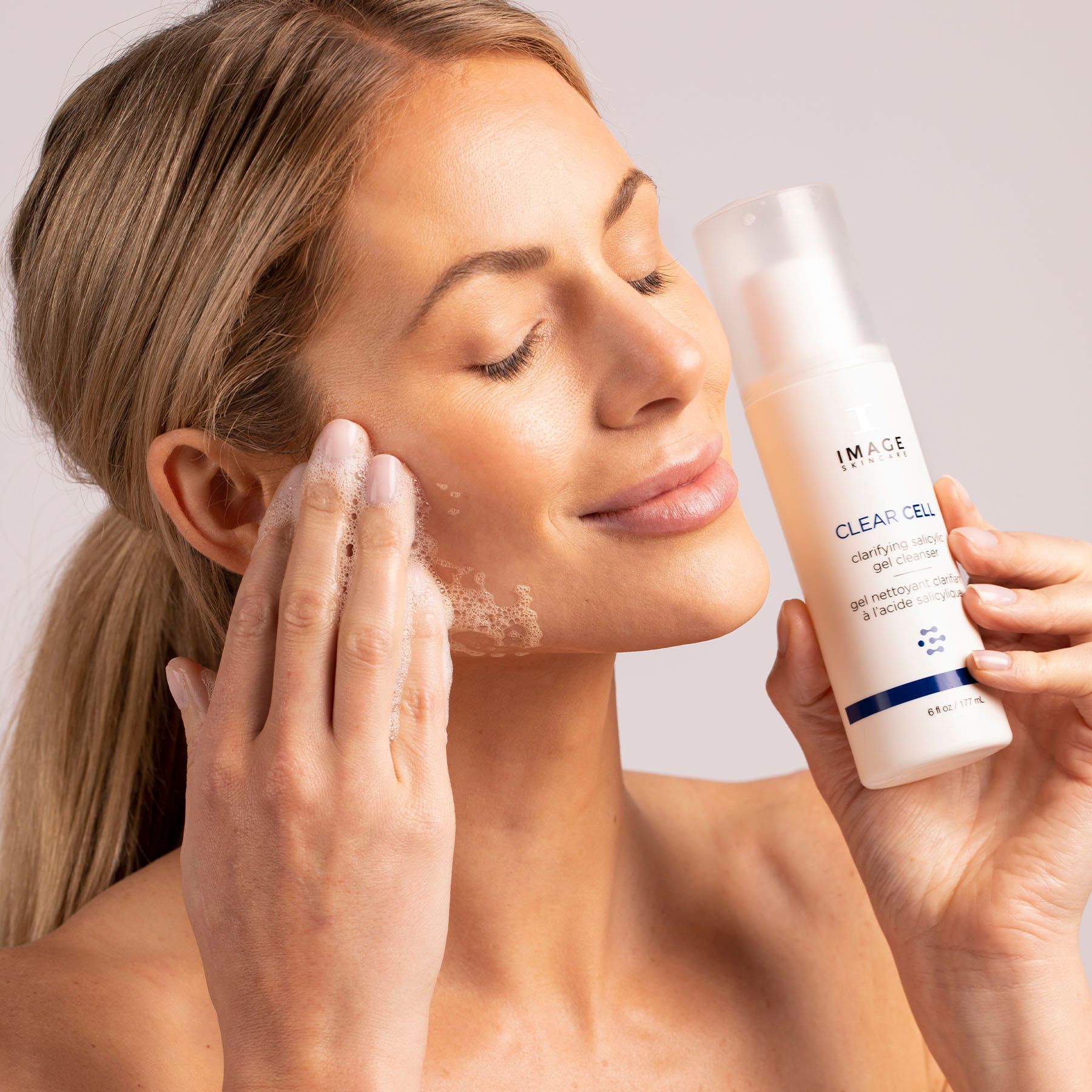 Woman applying face wash, eyes closed, holding bottle, light skin tone.