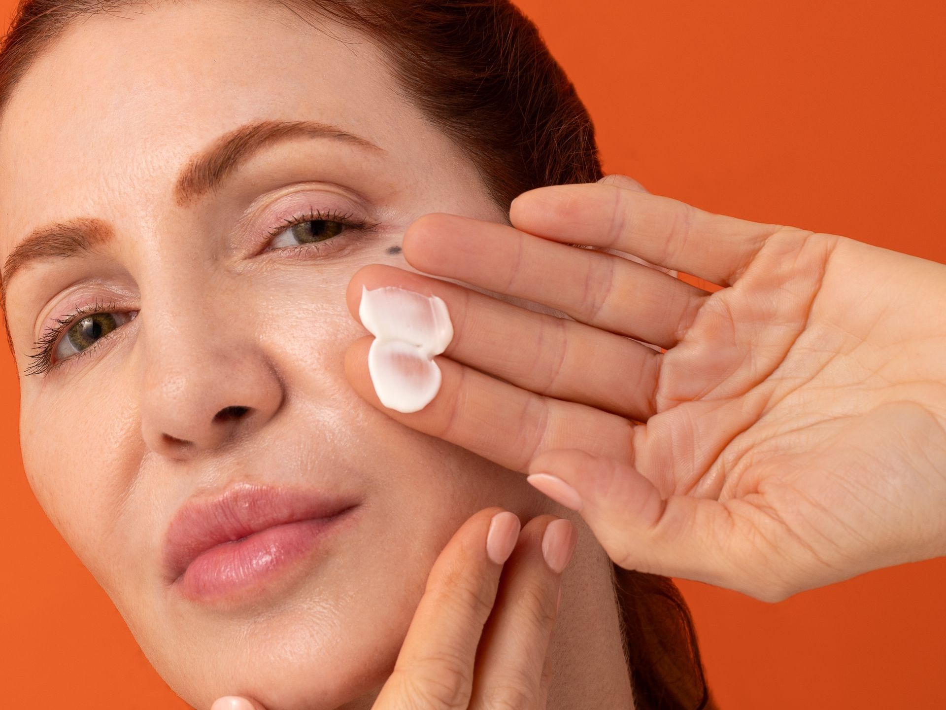 Woman applies cream to face with fingers; orange background.