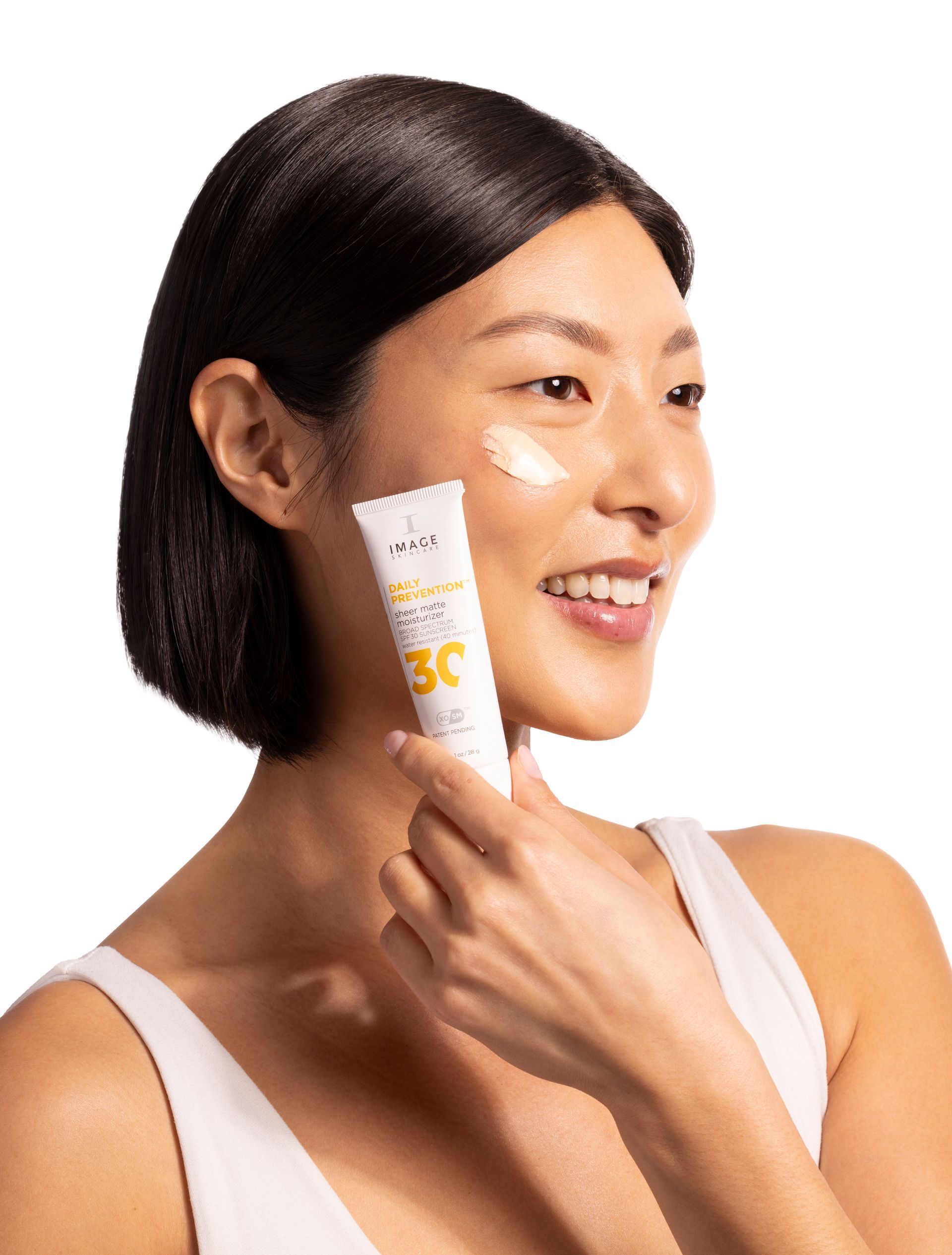 Woman smiling, applying sunscreen from a tube to her cheek. White background.