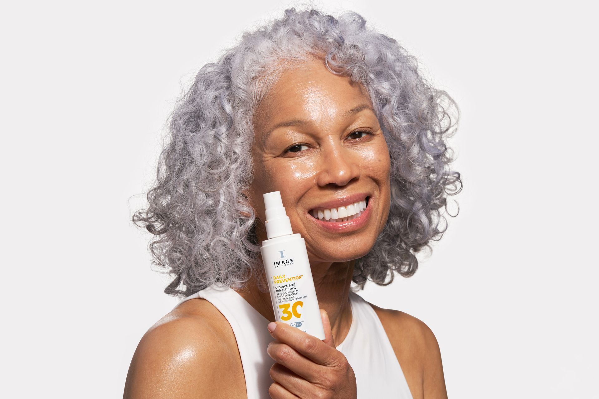 Smiling woman with gray curly hair, holding a sunscreen bottle, against a white background.