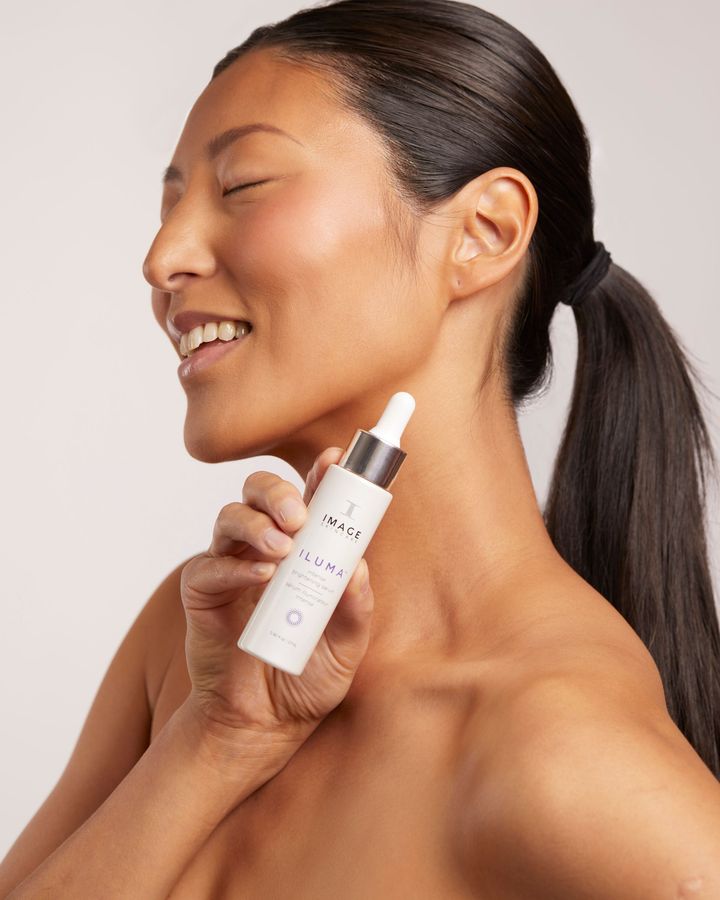 Woman smiling, holding skincare serum bottle, side view, ponytail, white background.