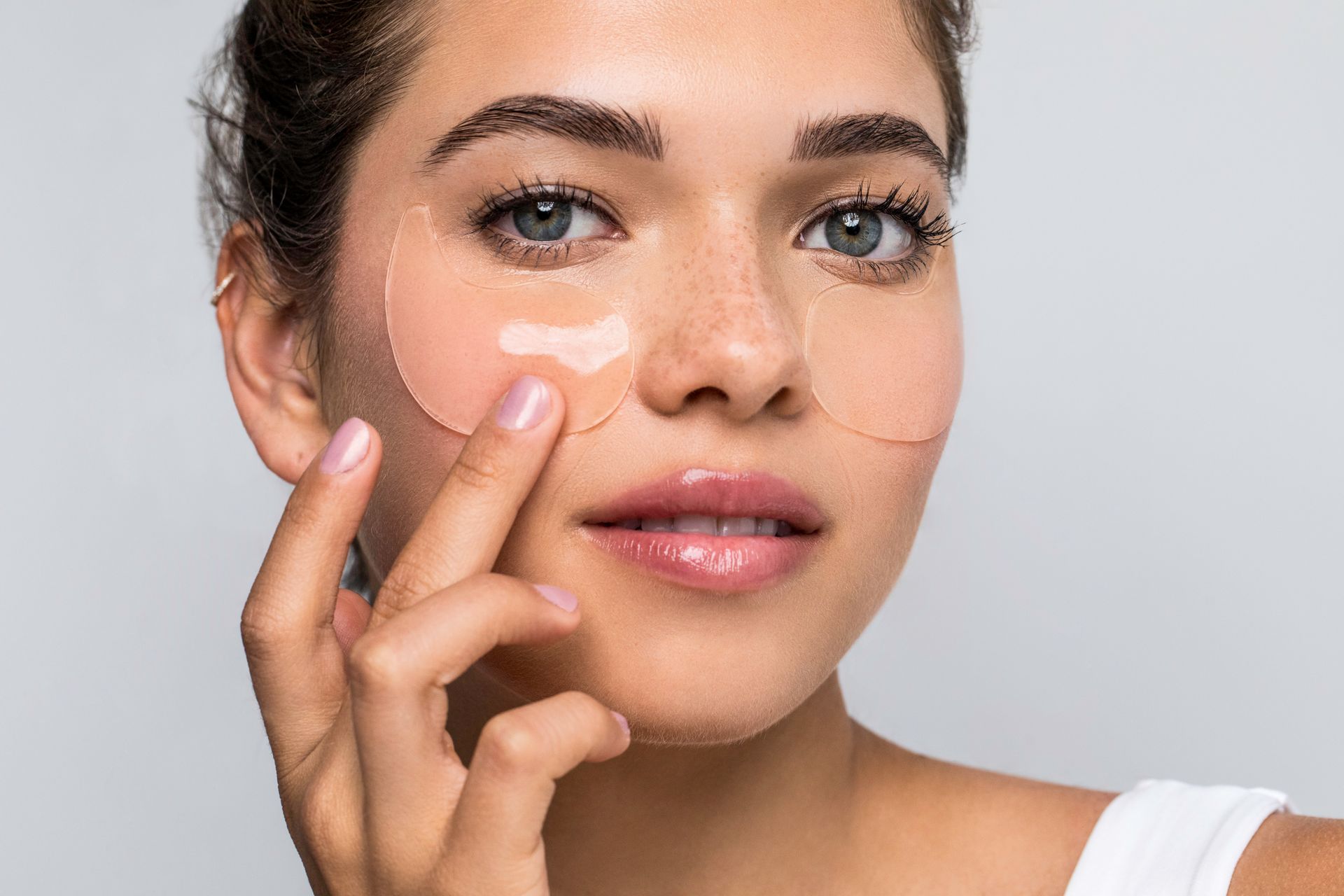 Woman applying under-eye patches. She has freckles and is touching her cheek with her fingers. Light background.