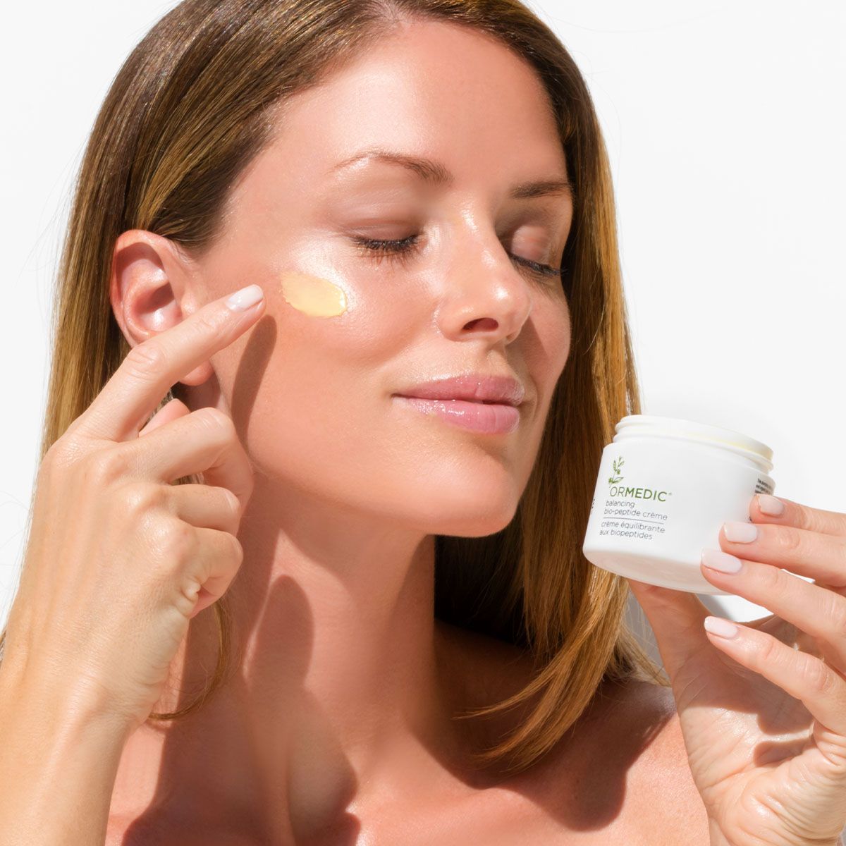 Woman applying skincare cream to her face, holding a white jar, eyes closed, light background.
