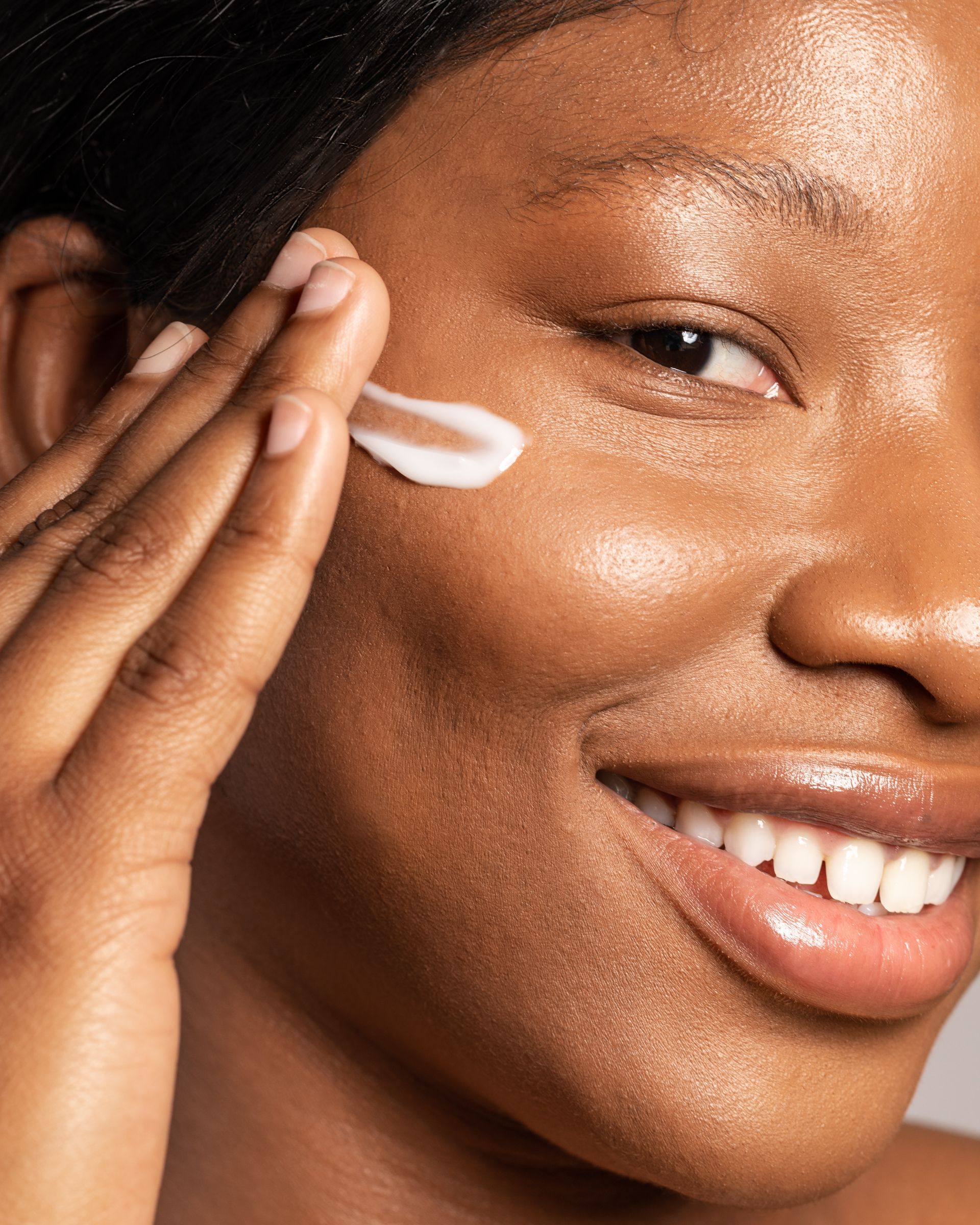 Woman with dark skin applying cream to her cheek, smiling.