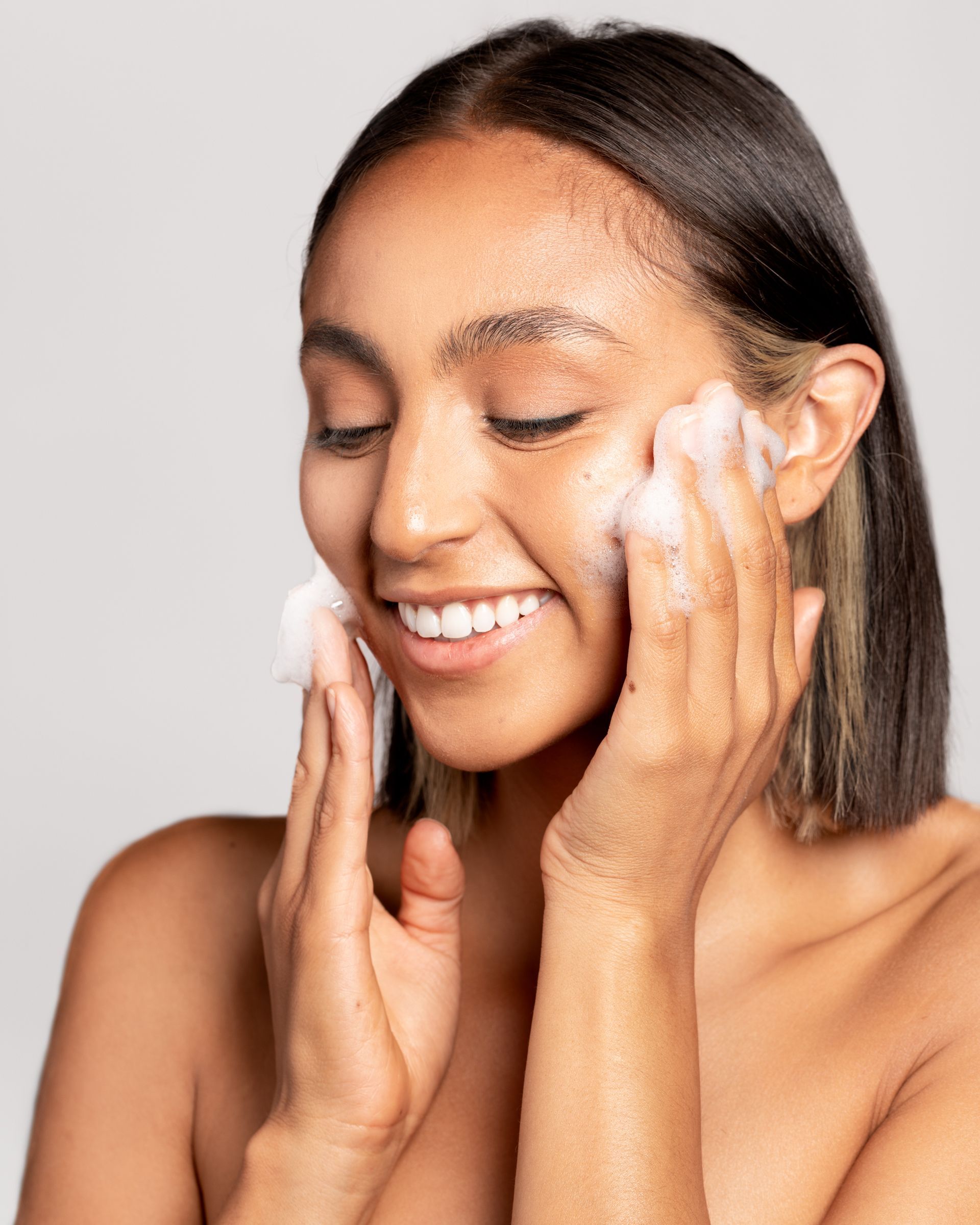 Woman washing face with white foam, smiling with eyes closed, studio setting.