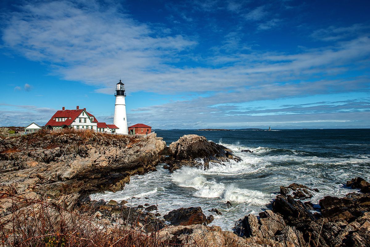 A lighthouse sits on top a rugged cliff, against a blue sky with ocean waves beating the shore.