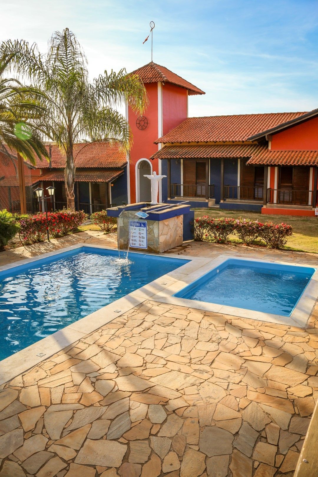 Cena externa com um prédio vermelho, piscina, pátio de pedra. Céu azul, ensolarado.