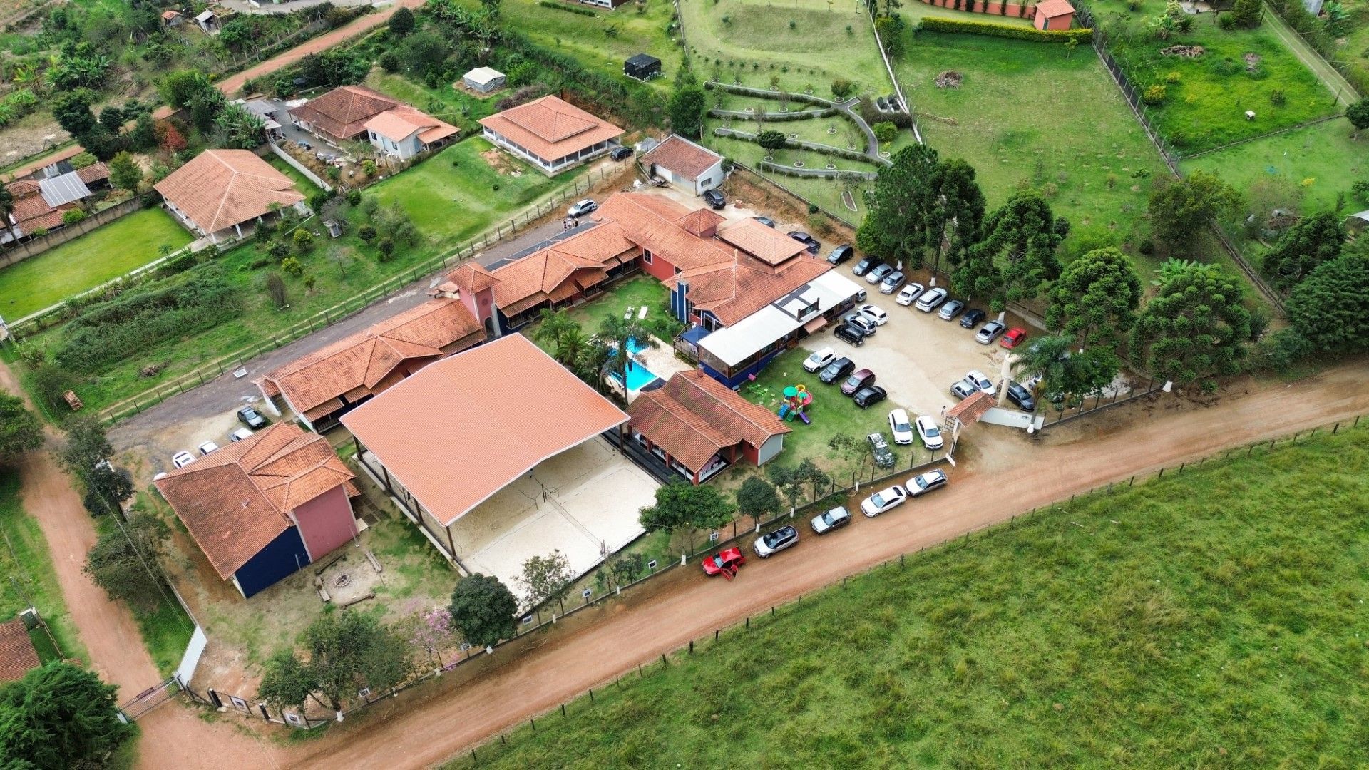 Vista aérea de um resort com telhados vermelhos, piscina, estacionamento e arredores verdes.