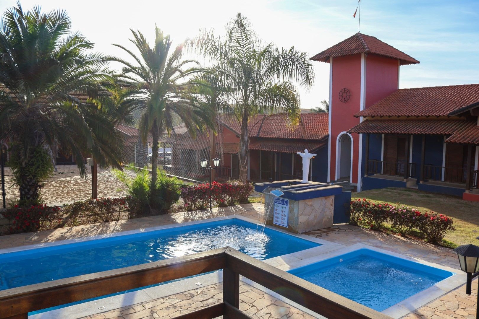 Piscina e prédio vermelho com palmeiras sob um céu ensolarado.